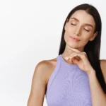 A detailed frontal view of a woman with long dark hair against a plain white background, wearing a lilac sleeveless top with her eyes closed and her index finger touching her chin to frame her jawline and submental profile for a Double Chin Liposuction in Dubai.