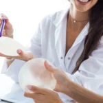 A close-up view of a smiling doctor in a white medical coat using a purple marker to point at a matte textured breast implant held by a patient, with a second glossy round implant held below it, evaluating What do implants look like after 20 years.