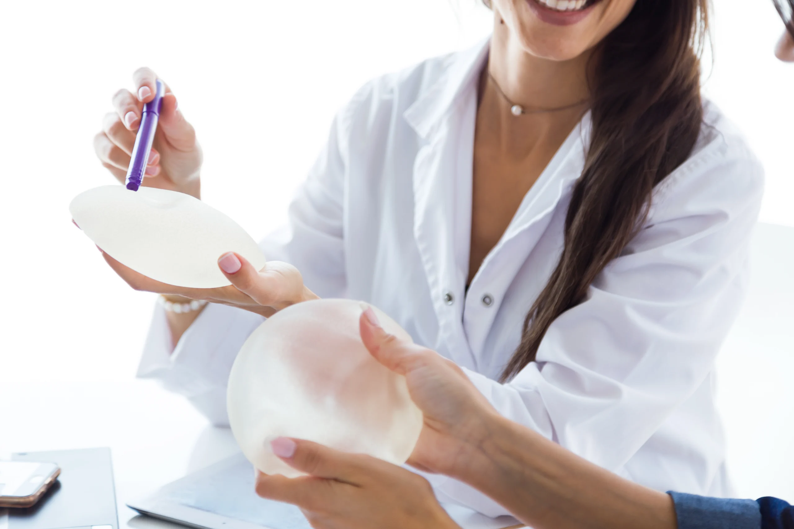 A close-up view of a smiling doctor in a white medical coat using a purple marker to point at a matte textured breast implant held by a patient, with a second glossy round implant held below it, evaluating What do implants look like after 20 years.