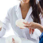 A close-up view of a surgeon in a white medical coat holding two textured mammary prosthetics while a patient points a finger at the upper gel shell to discuss indicators for When my breast implants need replacing.