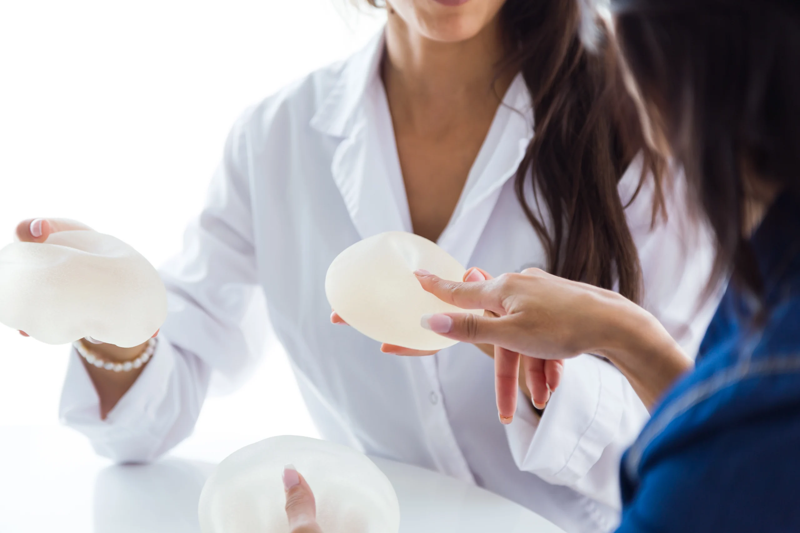 A close-up view of a surgeon in a white medical coat holding two textured mammary prosthetics while a patient points a finger at the upper gel shell to discuss indicators for When my breast implants need replacing.