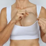 A close-up frontal view of a woman sliding a teardrop-shaped cohesive silicone implant inside her white sports bra to demonstrate the anatomical placement of a gummy bear breast implant.