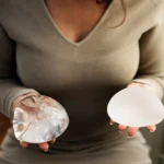 A close-up view of a woman wearing a grey long-sleeve dress, holding a clear, glossy silicone breast implant in her right hand and a matte, textured cohesive gel implant in her left hand to evaluate What happens if you never remove your breast implants.