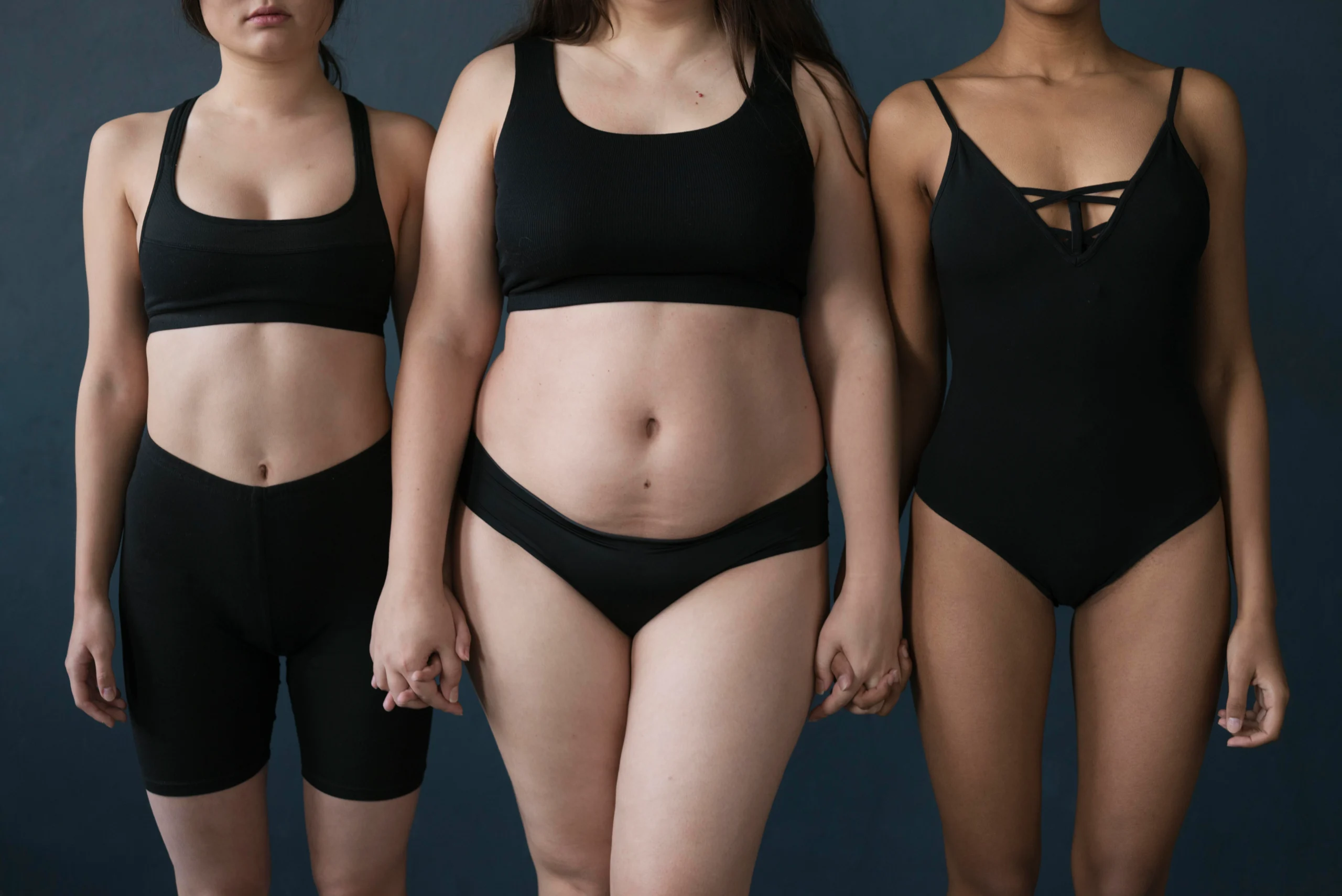 A frontal studio portrait of three women of diverse body types standing side-by-side in white underwear against a light beige background, showcasing varying abdominal profiles to illustrate the combined approach of Liposuction vs Tummy Tuck: Combined Procedures Dubai.