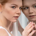 A female patient looking at her profile reflection in a mirror during an evaluation for Rhinoplasty Thin Skin vs Thick Skin in Dubai, focusing on the dorsal esthetic lines and nasal tip definition.