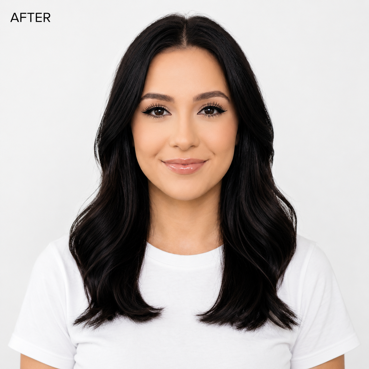 A frontal headshot of a smiling woman with styled, wavy black hair parted down the middle, wearing a white t-shirt and subtle makeup against a plain white background, featuring a text label reading "AFTER" in the upper-left corner to show the postoperative results of Blepharoplasty in Dubai.