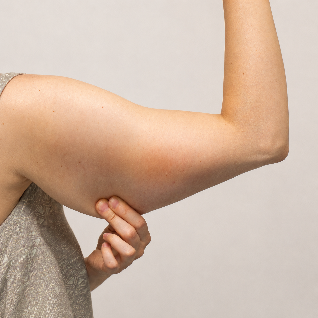 A close-up profile shot of a person's upper body against a light neutral background, showing an outstretched right arm bent at a 90-degree angle while a hand pinches the loose, sagging skin and excess fat hanging from the triceps region to demonstrate a baseline for an arm lift in Dubai.