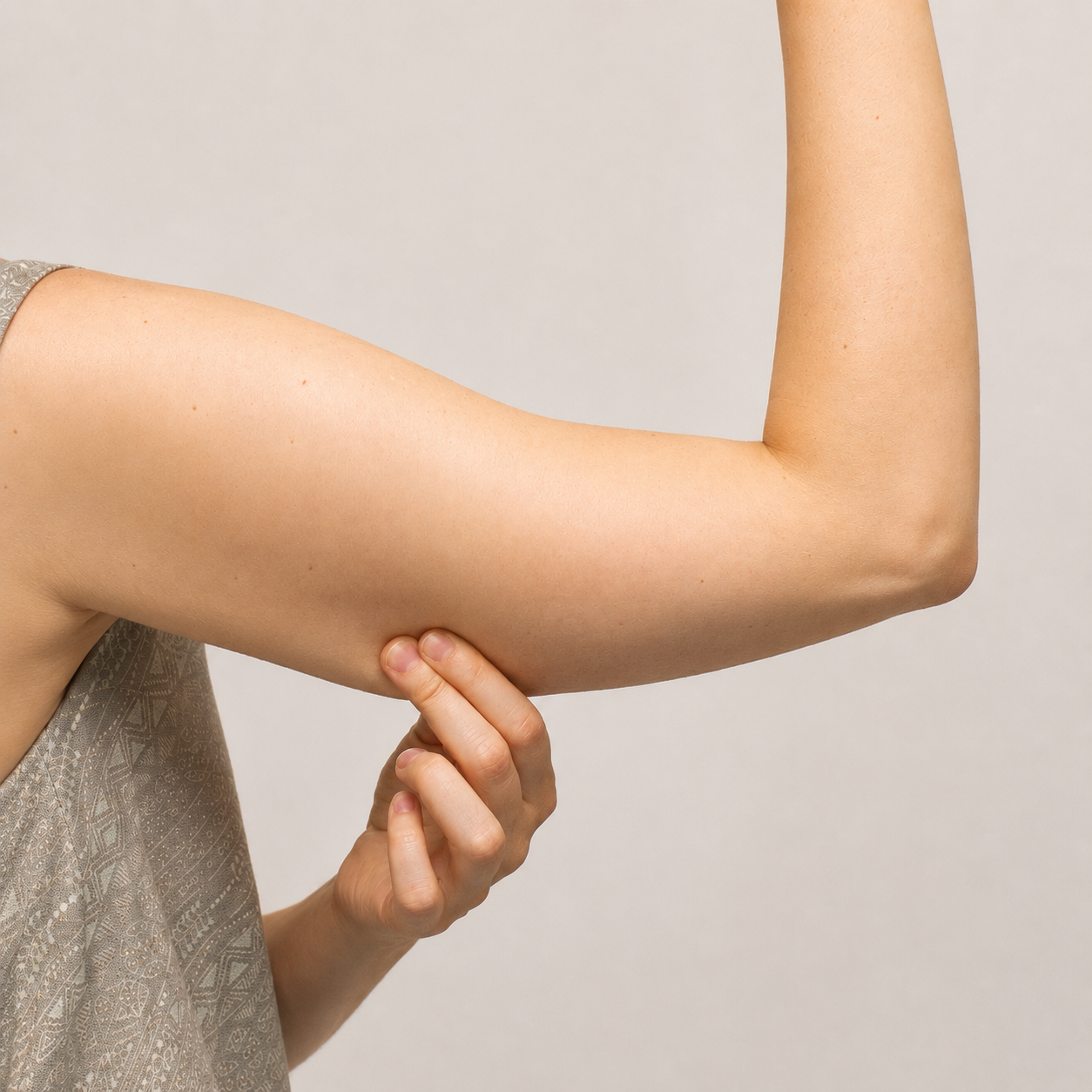 A close-up profile shot of a person's upper body against a light neutral background, showing a contoured right arm bent at a 90-degree angle while a hand holds the significantly firmed and reduced skin tissue along the triceps area to demonstrate the postoperative result of an arm lift in Dubai.