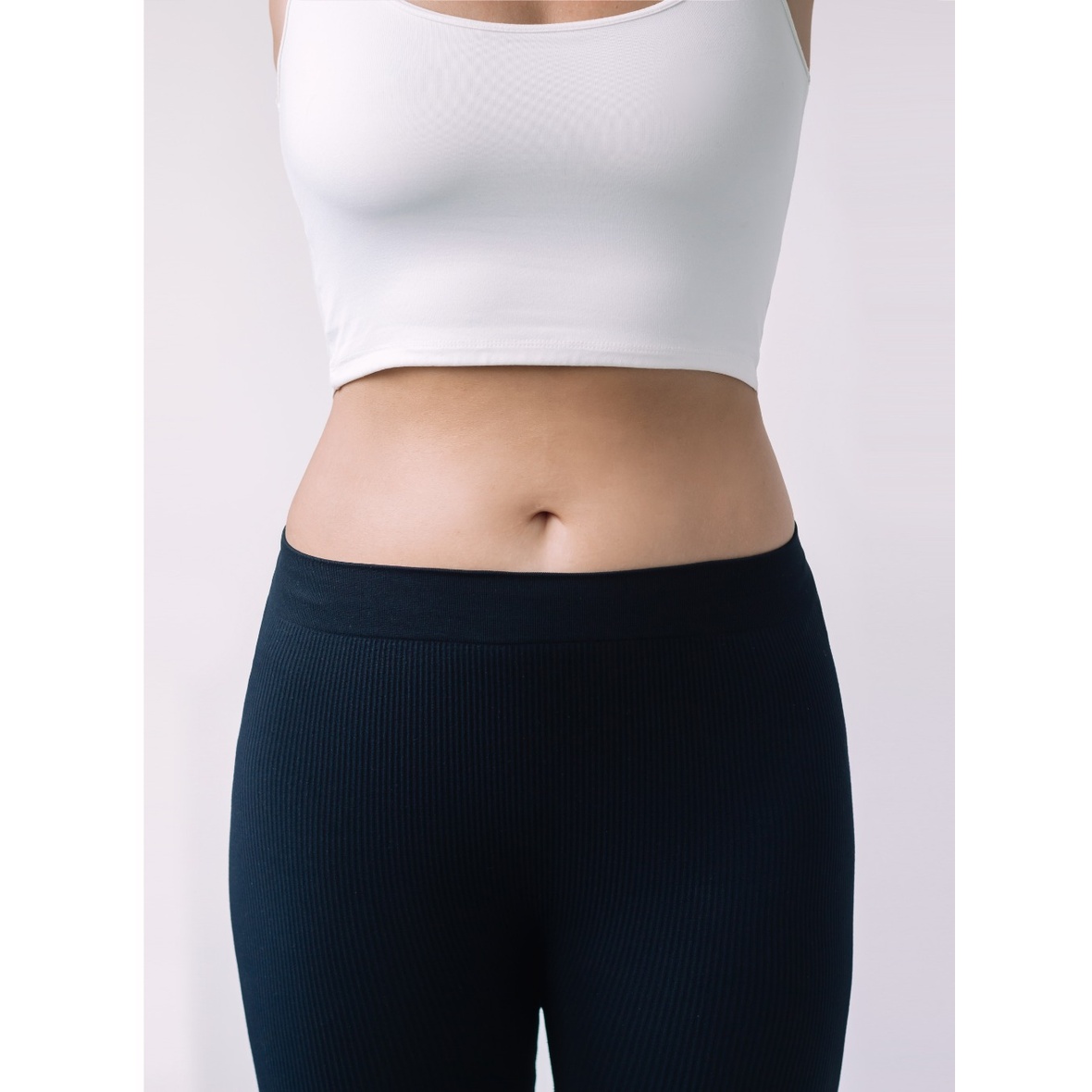 A frontal close-up shot from the chest to the upper thighs of a woman standing in a white crop top tank and dark ribbed athletic pants against a plain white background, showcasing a flat, highly contoured stomach and tapered waistline achieved after a Lipo Abdominoplasty in Dubai.