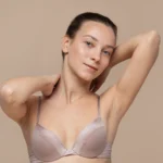 A detailed close-up frontal view of a smiling woman with dark hair tied back against a solid beige background, wearing a taupe-colored underwire bra with her arms raised behind her neck to display upper body contours and chest symmetry for a Hybrid Breast Augmentation Dubai.