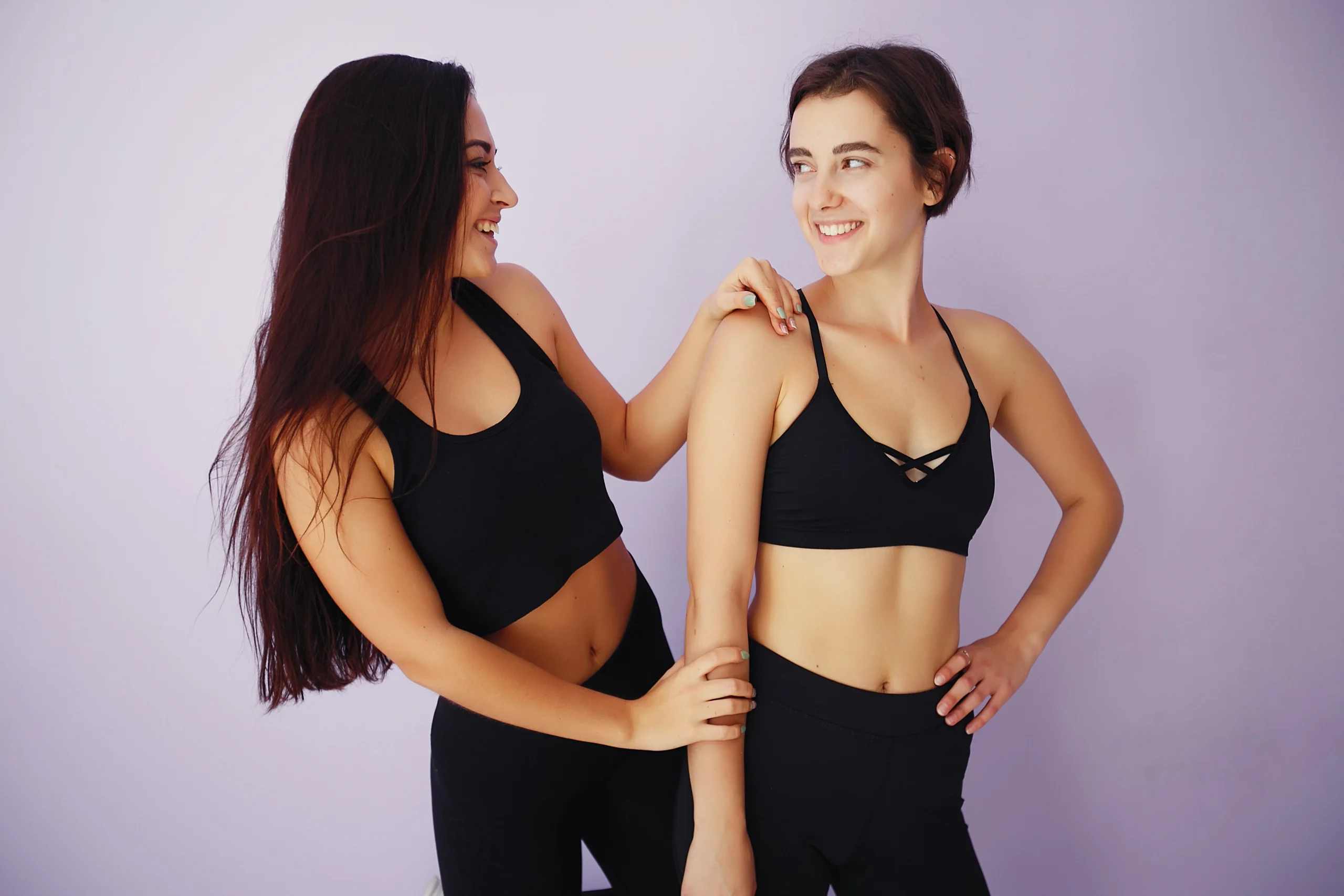A medium frontal shot of two young women in black sports bras and leggings standing side-by-side against a light purple background, smiling at each other and showcasing their toned abdominal muscles to compare High-definition liposuction vs liposuction.