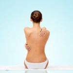 A full rear view of a topless woman with her hair in a bun sitting on a white towel in white panties against a light blue gradient background, hugging her shoulders to highlight her posterior contours and candidates for Back Liposuction in Dubai: Say Goodbye to Bra Fat and Achieve a Smoother Silhouette.