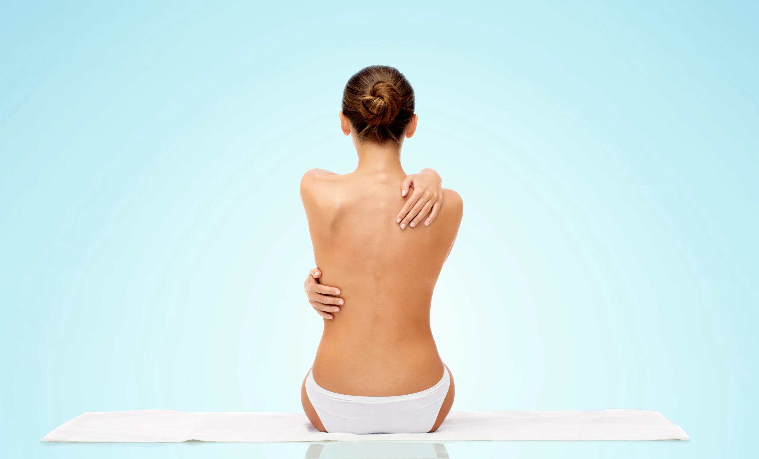 A full rear view of a topless woman with her hair in a bun sitting on a white towel in white panties against a light blue gradient background, hugging her shoulders to highlight her posterior contours and candidates for Back Liposuction in Dubai: Say Goodbye to Bra Fat and Achieve a Smoother Silhouette.
