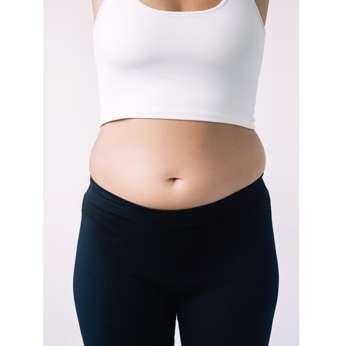 A frontal close-up shot from the chest to the upper thighs of a woman standing in a white crop top tank and dark ribbed athletic pants against a plain white background, showcasing localized belly fat and waist fullness to establish the baseline for a Lipo Abdominoplasty in Dubai.