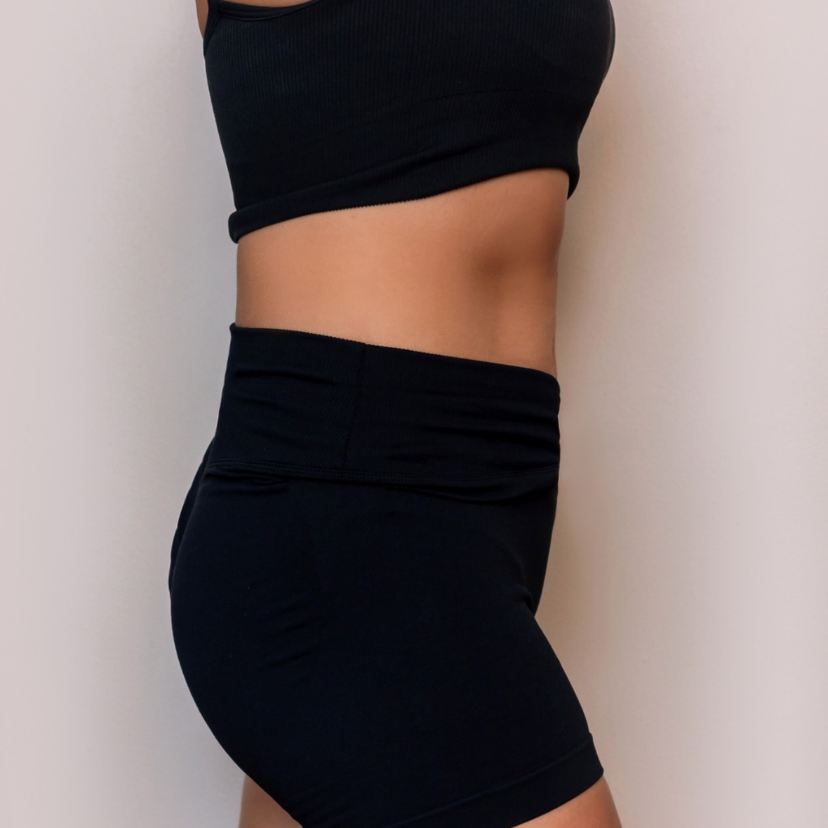 A side profile close-up shot from the chest to the upper thighs of a woman standing in a black sports bra and matching black athletic shorts against a plain light background, showcasing a flat, tightened stomach profile and streamlined waistline achieved after Waist Liposuction in Dubai.