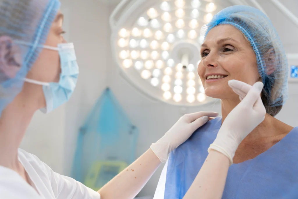 An over-the-shoulder view of a surgeon in a face mask and hairnet examining a smiling female patient under a bright operating room light, gently touching her chin and neck area to assess structural profile alignment for Rhinoplasty Tip Reshaping.