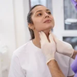 A professional front-facing clinical view of a woman sitting in a medical room with her chin slightly elevated while a surgeon wearing purple scrubs and sterile white gloves gently palpates her anterior neck tissue to evaluate underlying muscle structures for a Platysmaplasty Dubai.
