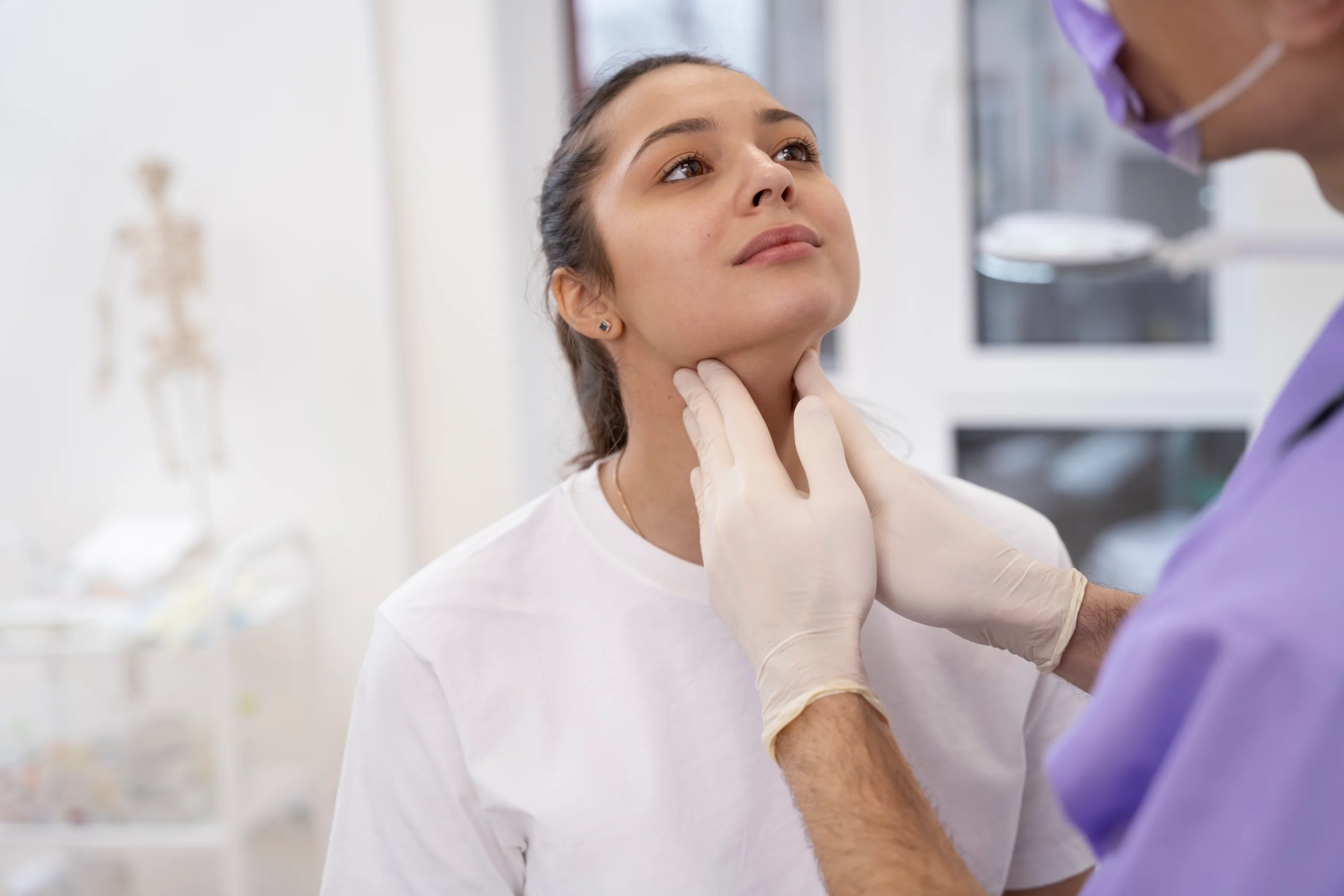 A professional front-facing clinical view of a woman sitting in a medical room with her chin slightly elevated while a surgeon wearing purple scrubs and sterile white gloves gently palpates her anterior neck tissue to evaluate underlying muscle structures for a Platysmaplasty Dubai.