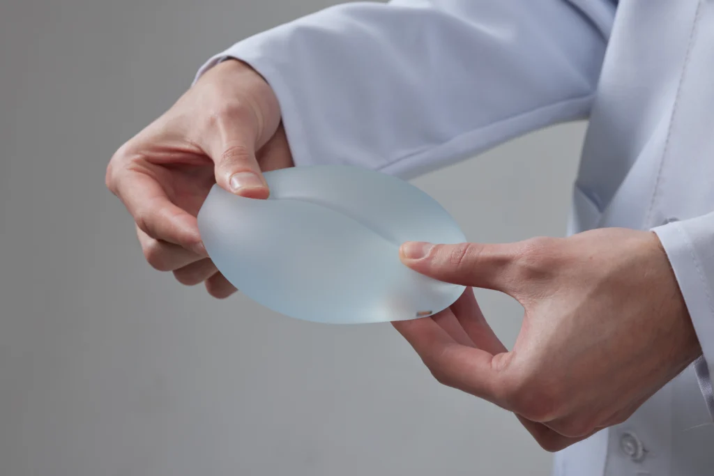 A detailed close-up view of a surgeon’s hands in a white medical coat gently pressing and flexing a textured silicone gel breast prosthesis against a neutral gray background to demonstrate material durability and shell flexibility for a Breast Implant in dubai.
