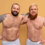 A front view of two shirtless, smiling men standing side-by-side in grey underwear against a warm yellow background, showcasing their natural abdominal outlines and chest framing to evaluate parameters for HD Lipo Sculpture In Dubai.
