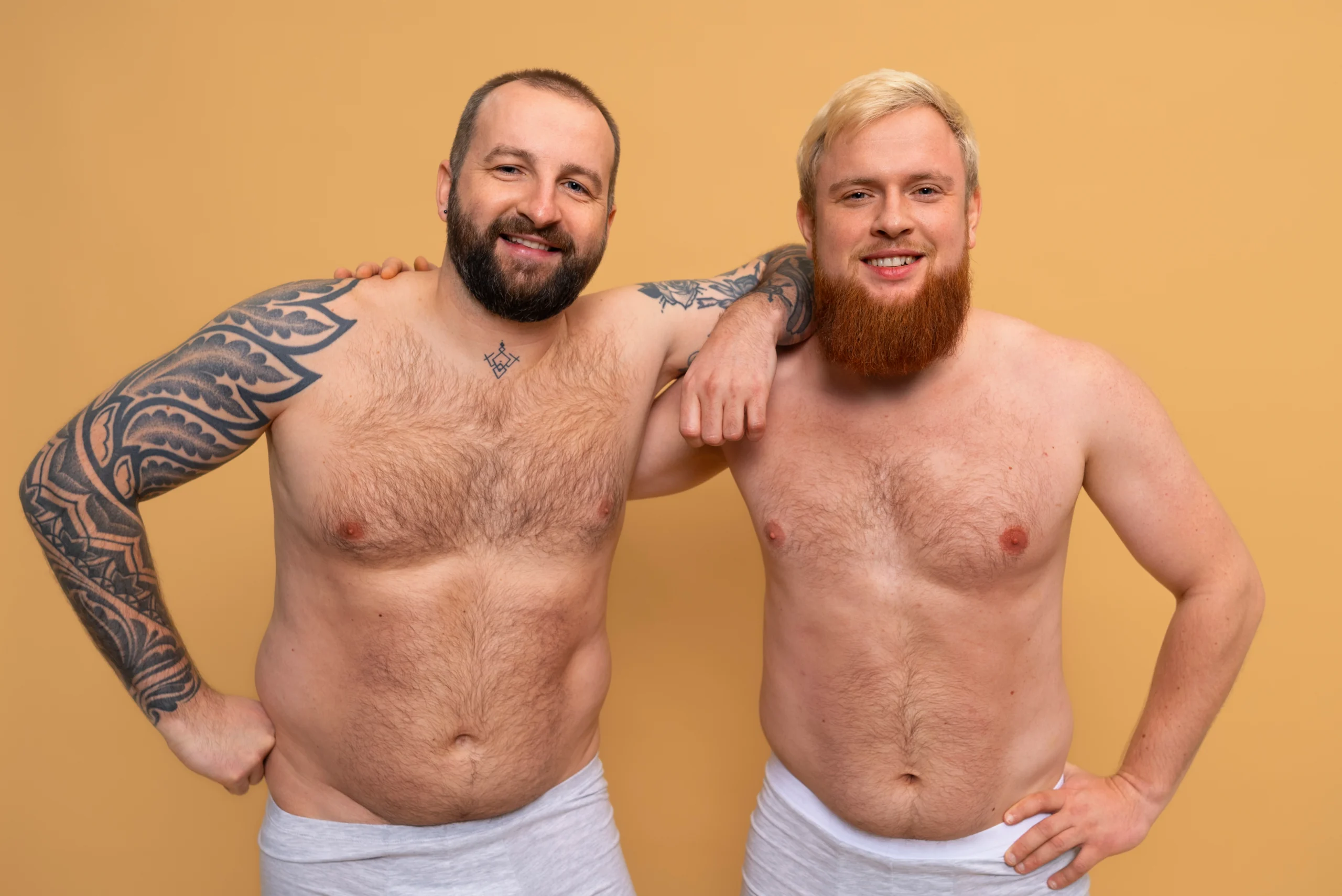 A front view of two shirtless, smiling men standing side-by-side in grey underwear against a warm yellow background, showcasing their natural abdominal outlines and chest framing to evaluate parameters for HD Lipo Sculpture In Dubai.