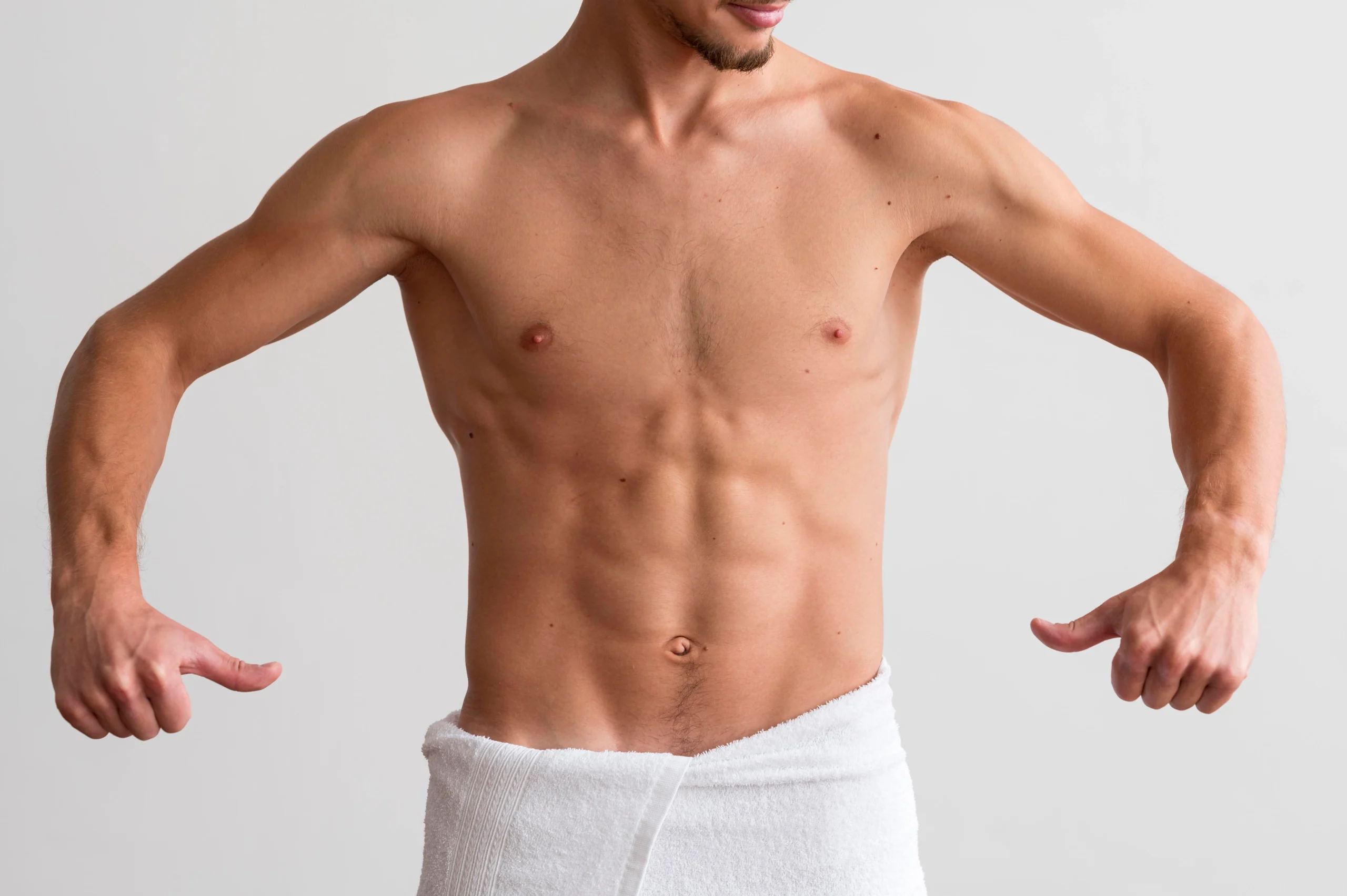 A frontal view of a shirtless male patient wrapped in a white towel, pointing toward his highly contoured torso to demonstrate the abdominal wall definition achieved through V Sculpt Liposuction Dubai.