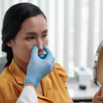 A detailed three-quarters clinical view of a woman with dark hair tied back looking into a hand mirror, while a plastic surgeon wearing blue sterile gloves gently examines her nasal tip and dorsum to plan a Revision Rhinoplasty Dubai.
