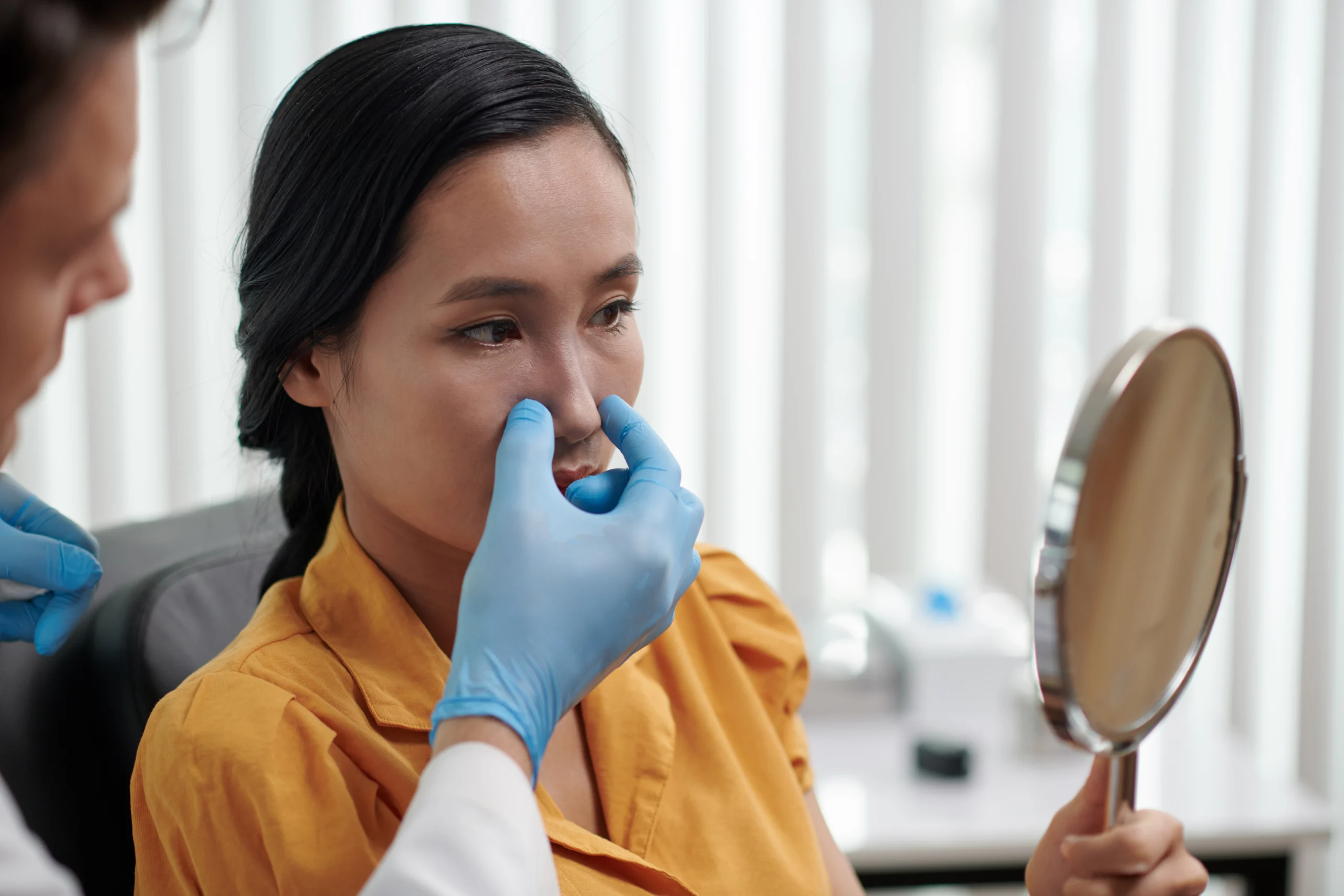 A detailed three-quarters clinical view of a woman with dark hair tied back looking into a hand mirror, while a plastic surgeon wearing blue sterile gloves gently examines her nasal tip and dorsum to plan a Revision Rhinoplasty Dubai.