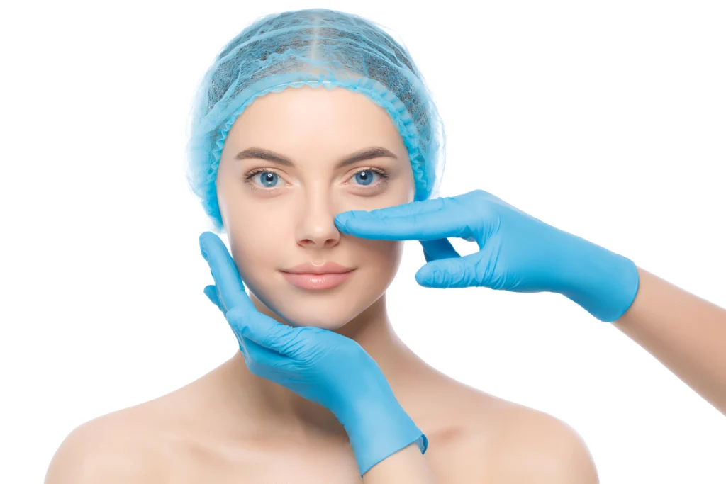 A frontal studio portrait of a young woman wearing a light blue surgical hairnet against a white background, while a surgeon's hands in blue nitrile gloves examine her nasal structure, placing one hand along her jawline and an index finger gently touching the side of her nose to evaluate eligibility for Rhinoplasty Tip Reshaping.