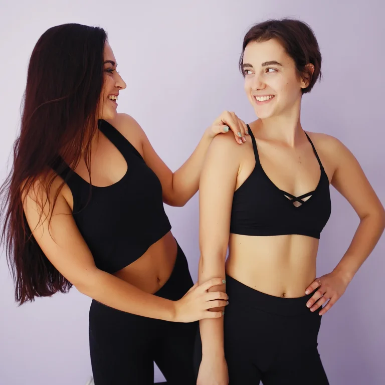 A medium frontal shot of two young women in black sports bras and leggings standing side-by-side against a light purple background, smiling at each other and showcasing their toned abdominal muscles to compare High-definition liposuction vs liposuction.