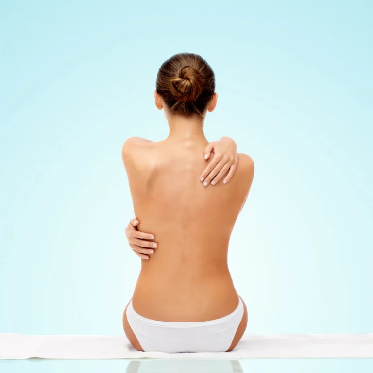 A full rear view of a topless woman with her hair in a bun sitting on a white towel in white panties against a light blue gradient background, hugging her shoulders to highlight her posterior contours and candidates for Back Liposuction in Dubai: Say Goodbye to Bra Fat and Achieve a Smoother Silhouette.