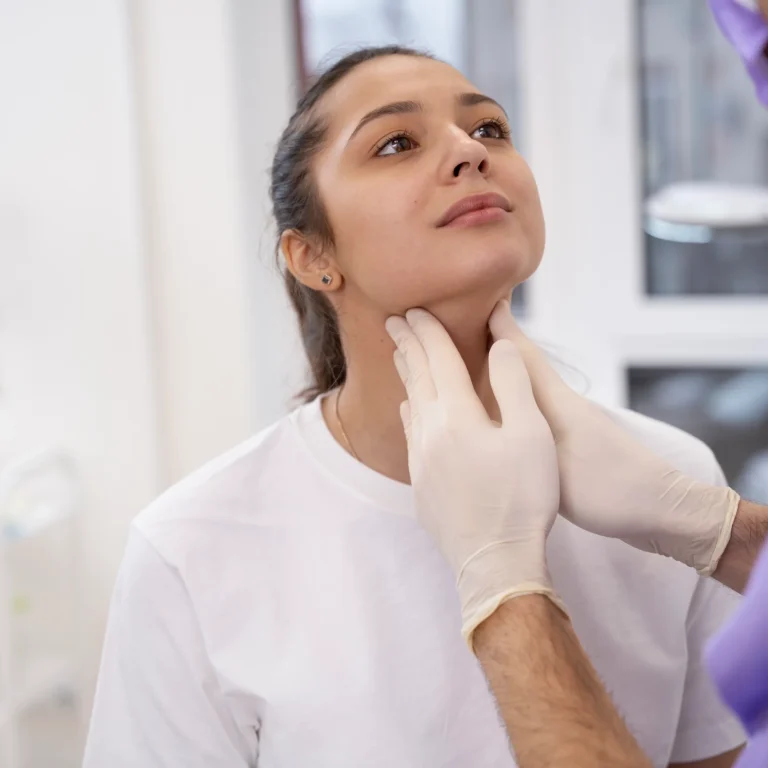 A professional front-facing clinical view of a woman sitting in a medical room with her chin slightly elevated while a surgeon wearing purple scrubs and sterile white gloves gently palpates her anterior neck tissue to evaluate underlying muscle structures for a Platysmaplasty Dubai.