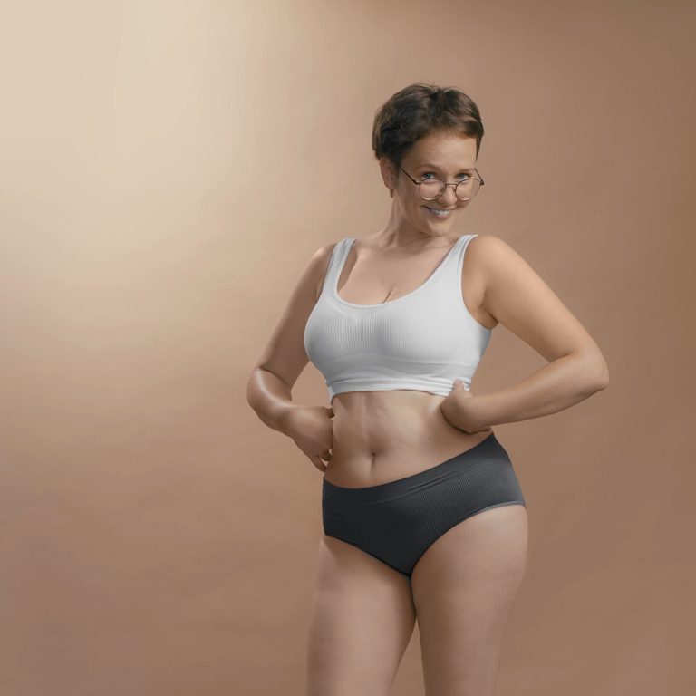 Chubby female in white bra and black briefs smiles, looks to the camera and adores her forms. Hands on the waist. Studio cowboy shot photo image.