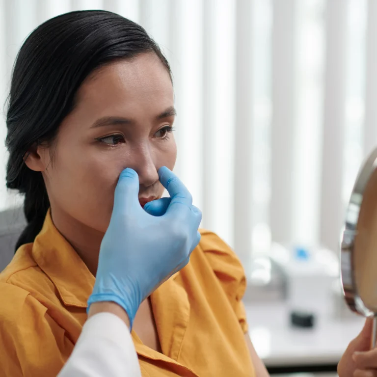 A detailed three-quarters clinical view of a woman with dark hair tied back looking into a hand mirror, while a plastic surgeon wearing blue sterile gloves gently examines her nasal tip and dorsum to plan a Revision Rhinoplasty Dubai.