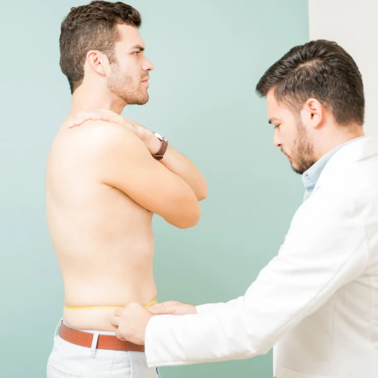 A plastic surgeon in a white coat conducting a pre-operative evaluation by measuring a male patient's waistline with a tape measure, highlighting why V Sculpt Liposuction is safe for men over 50 during personalized clinical assessments.